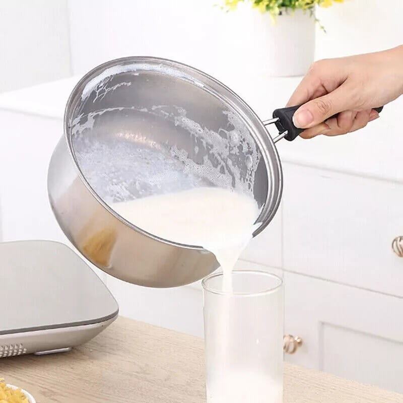 Se muestra un cazo de acero inoxidable con asa negra de un juego de utensilios de cocina de acero inoxidable para inducción con tapas de vidrio vertiendo leche en un vaso.