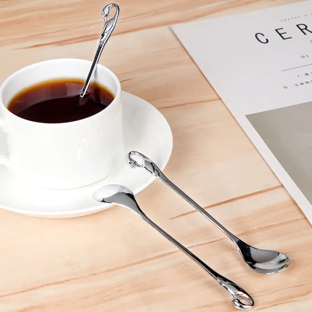 Une paire de cuillères à dessert métalliques élégantes à tons argentés, dotées de poignées torsadées ornées, reposant à côté d’une tasse à café en céramique blanche, du Coffret Swan Cutlery Set Gold.