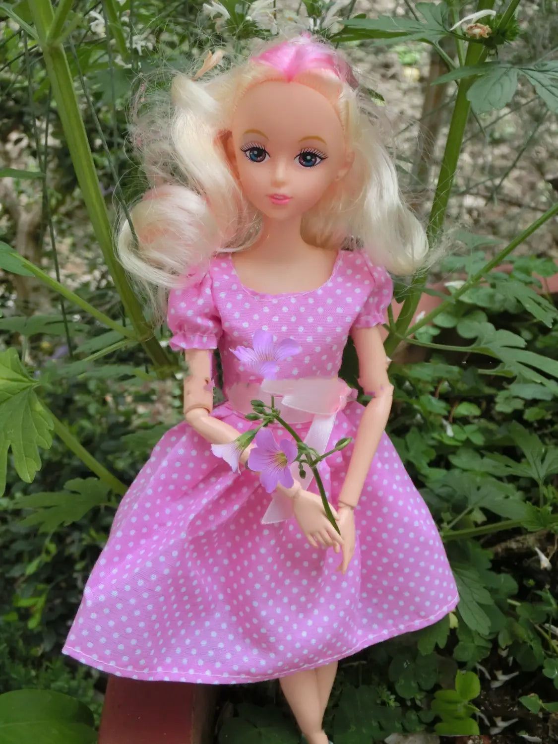 Eine blonde Anziehpuppe mit langem welligem Haar in einem pinkfarbenen Fashion-Set mit Punkten, die für Kinder lila Blumen hält.