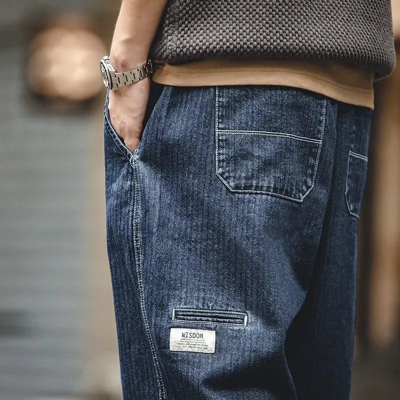 Pantalones vaqueros relajados y cónicos azul marino oscuro para hombre con lavado deslavado, costuras blancas y un parche rectangular de la marca Wisdom en la pierna inferior.