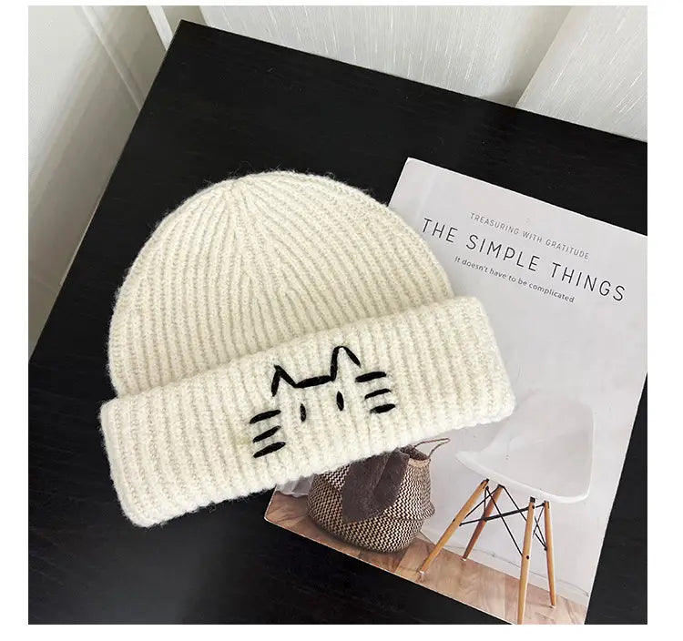 Cream-colored knitted beanie hat with a black embroidered cat face on the folded brim.