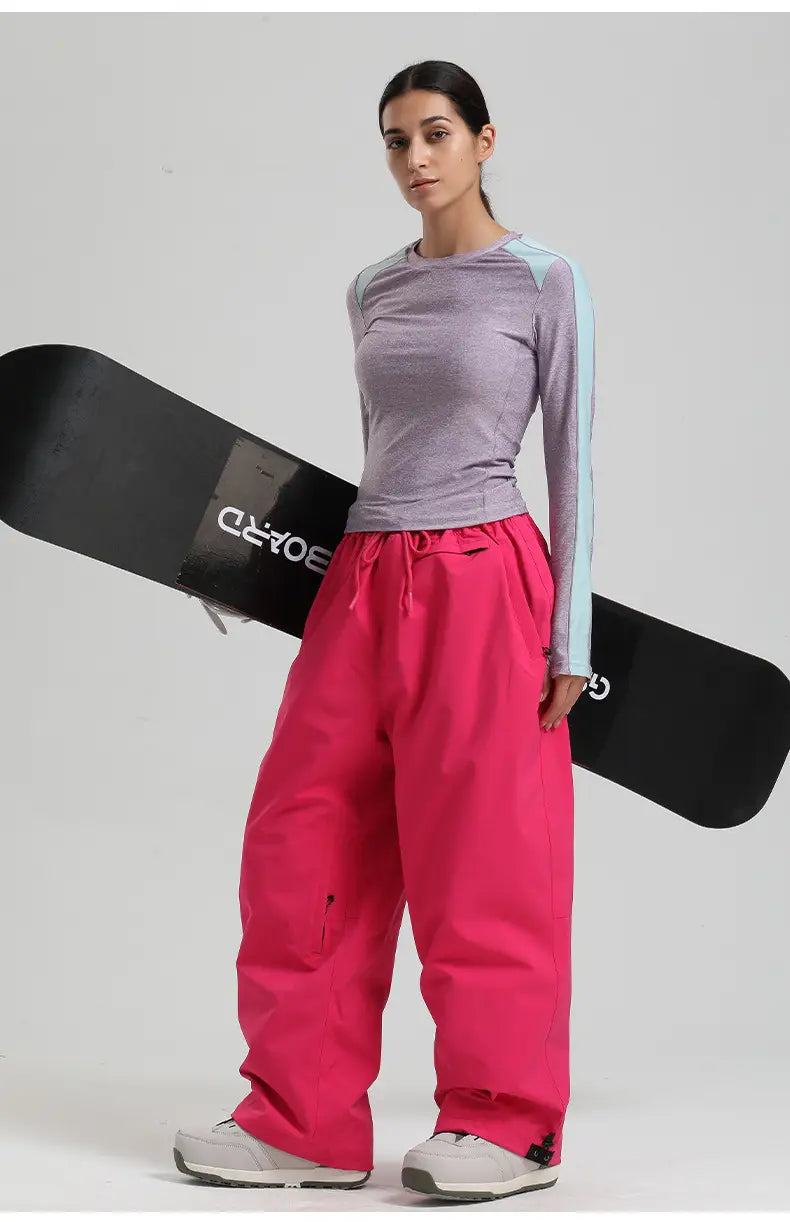 a model wearing vibrant pink waterproof windproof snow pants with a drawstring waist and side pockets holding a black snowboard.