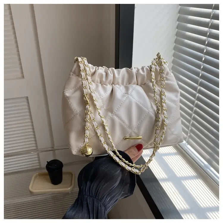Cream-colored quilted faux leather crossbody bag with satin finish gold-tone chain straps and decorative golden ball charm.