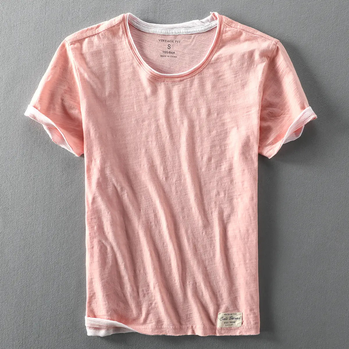 Ein helles rosafarbenes Herren-T-Shirt aus Baumwolle mit Kontrastbesatz, mit subtil gewebter Struktur und weißem Besatz entlang des Halsausschnitts, der Ärmel und des Saums, ideal für Freizeitkleidung im Sommer.