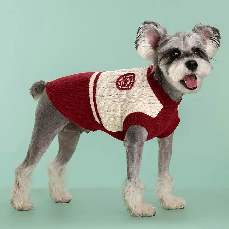 Ein gemütlicher roter und cremefarbener Zopfmuster-Hundepullover aus warmem Strickmaterial mit weißen Streifen an den Ärmeln und einem runden bestickten Aufnäher, geeignet für Tierbekleidung.