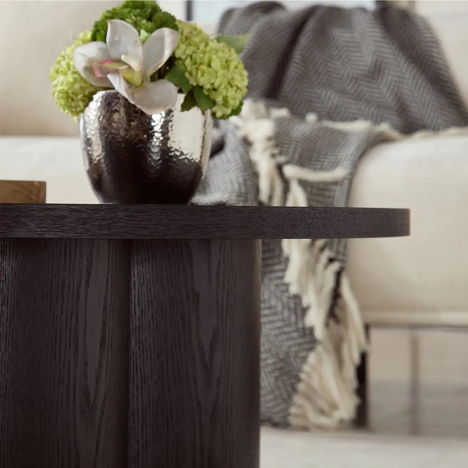 Mesa de centro redonda de madera oscura con un acento esculpido moderno que sostiene un florero metálico brillante con acabado texturizado tipo martillado, que contiene hortensias verdes y orquídeas blancas.