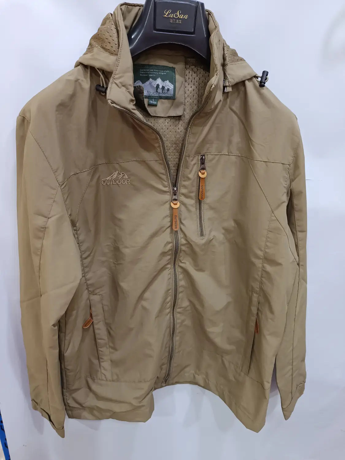 Chaqueta de senderismo con capucha impermeable color camel, con cierre frontal mediante cremallera y dos bolsillos laterales, con tiradores de cuero.