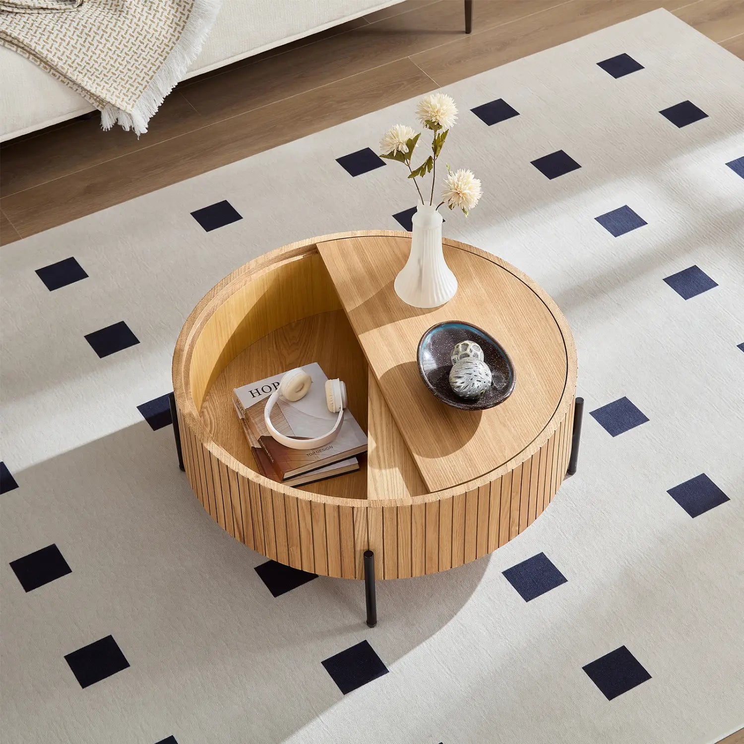 Mesa auxiliar redonda con almacenamiento: presenta un detalle de madera con lamas, patas de metal negras y dos compartimentos para guardar libros, auriculares y un jarrón blanco con flores.