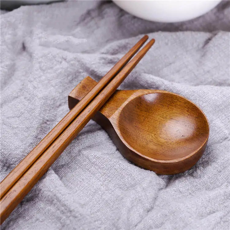 Un plato para salsa de inmersión de madera de color marrón medio y acabado liso, con soporte para palillos integrado, que sostiene un par a juego de palillos de madera.