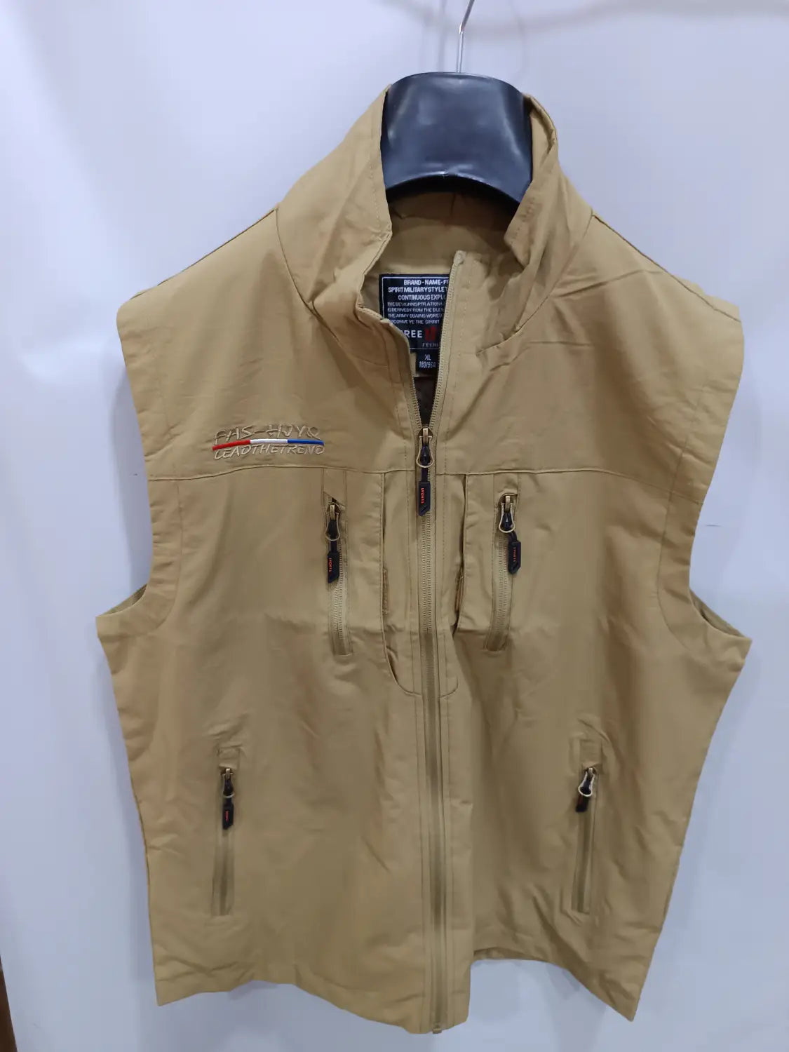Chaleco de pesca impermeable color arena para hombre con varios bolsillos con cremallera y un logotipo de la marca bordado en el lado izquierdo del pecho, diseñado como un chaleco utilitario con capucha y múltiples bolsillos.