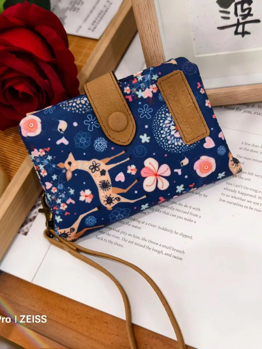 A navy blue floral-patterned canvas tri fold multi card wallet with tan leather accents and a matching strap.