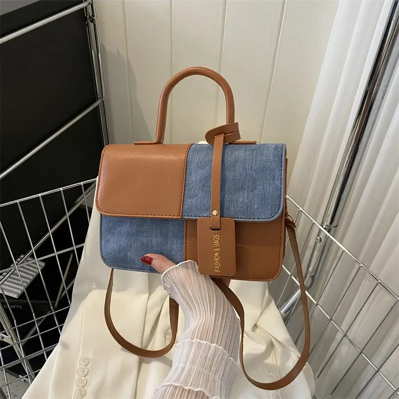 A two tone denim patchwork crossbody bag featuring a tan leather top flap and blue denim panel with matching brown straps.