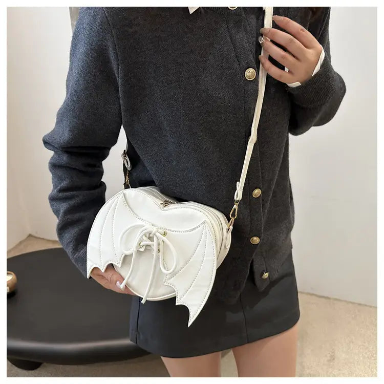 White bat-shaped crossbody bag featuring lace-up front detail and gold-tone hardware called Bat Wing Heart Crossbody Bag with Lace Up Detail.