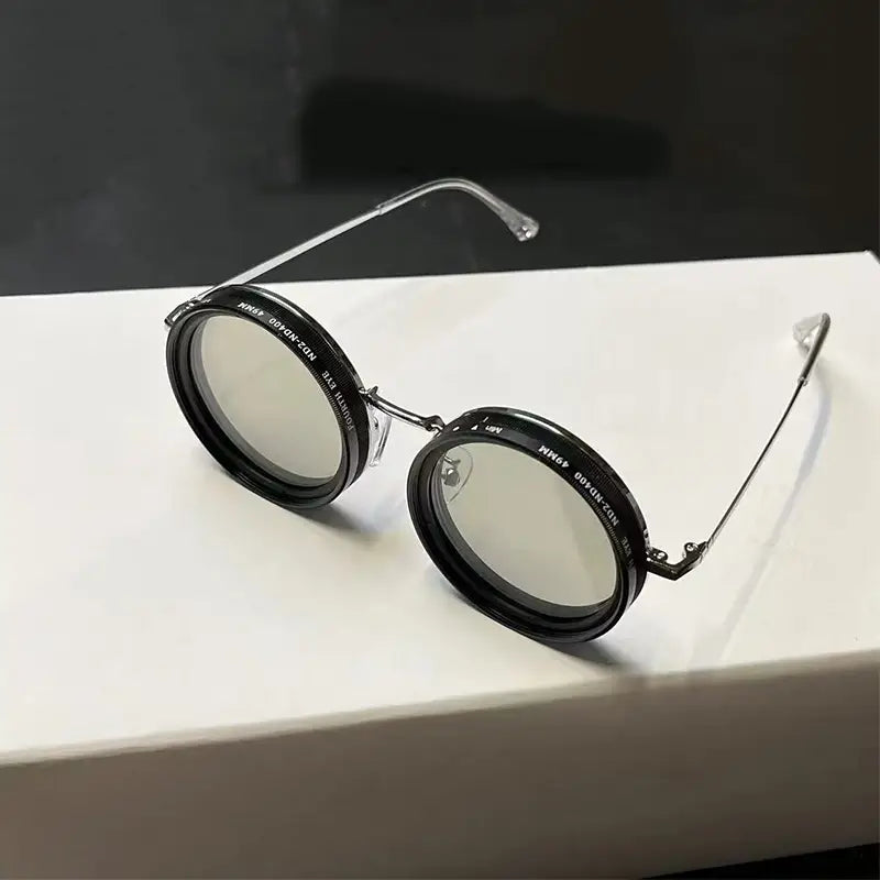 Lunettes de soleil rondes unisexes résistantes aux UV, avec montures circulaires noires, branches en métal à tons argentés et verres transparents.