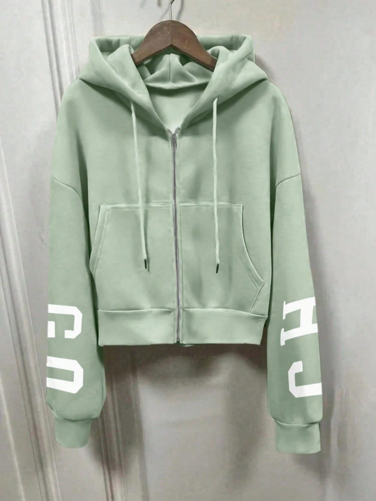 Sweat-shirt à capuche zippé imprimé graphique vert menthe court, avec lettrage blanc sur les manches, suspendu à un cintre en bois.