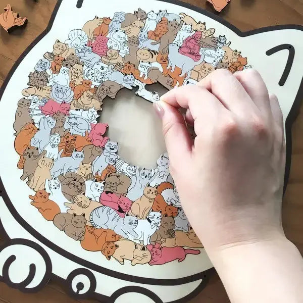 Una mano coloca una pieza de rompecabezas en un rompecabezas de madera con forma de gato de 135 piezas, tipo juguete rompecabezas Morandi, que incluye gatos ilustrados en varios colores y posturas sobre un fondo blanco con contornos negros.