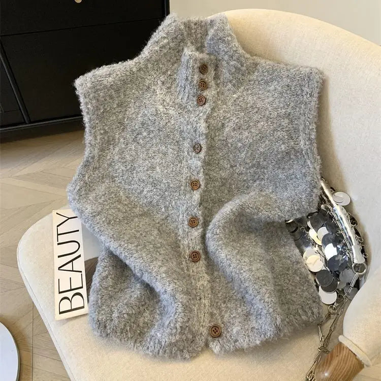 Un cardigan sans manches en maille gris clair duveteux et doux, avec un col montant et des boutons en bois bruns, évoquant un gilet tricoté à col haut, boutonné, crème.