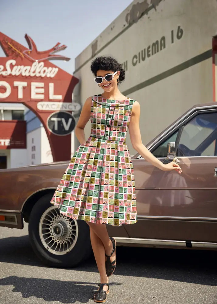 A woman wearing a sleeveless knee-length cotton a-line Elizabeth dress in vibrant vintage stamp print paradise colors