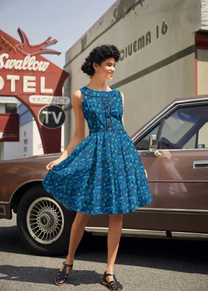 A woman wears an Elizabeth peacock print vintage cotton midi dress in blue with sleeveless design and black button detailing