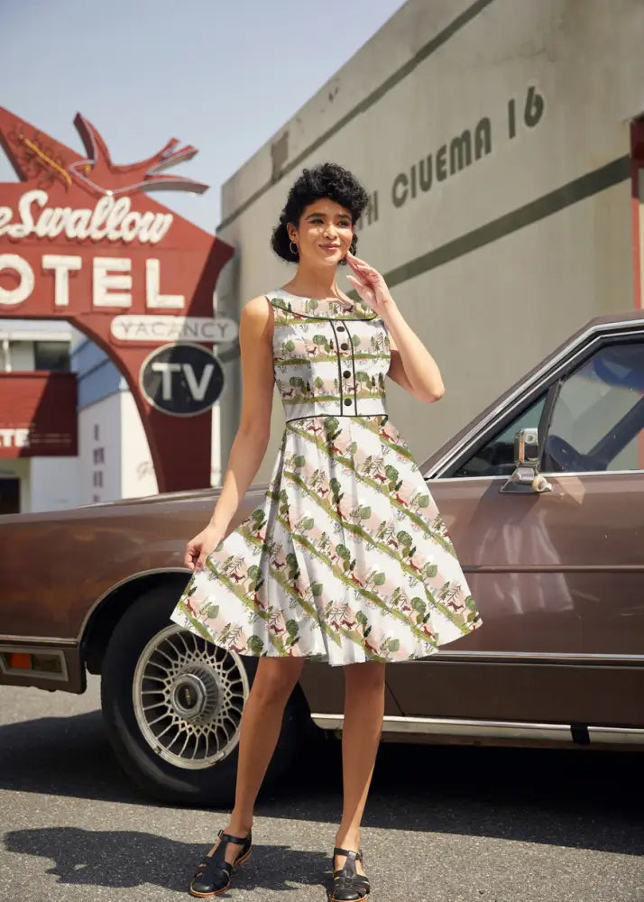 A woman wears the sleeveless knee-length Elizabeth Dress in white woodland sunset cotton print with green trees and brown animals