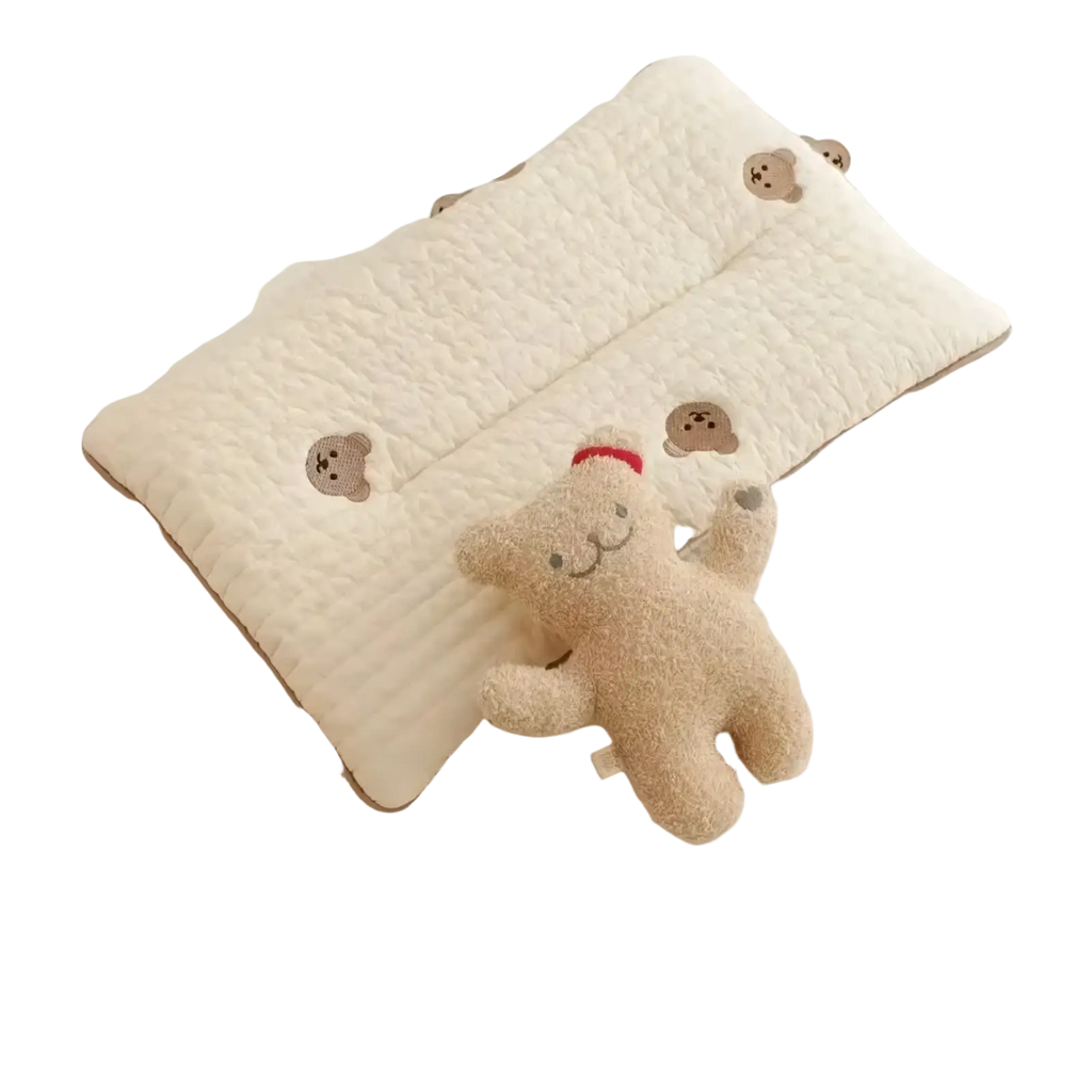 Cream-colored quilted embroidered cotton baby pillow with brown bear faces next to a plush beige teddy bear in red hat