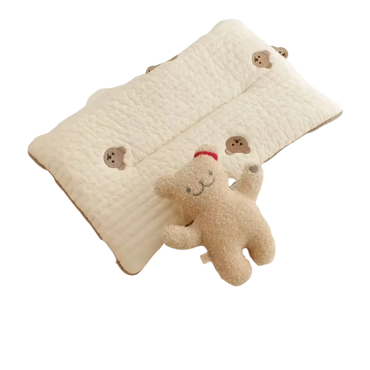 Cream-colored quilted embroidered cotton baby pillow with brown bear faces next to a plush beige teddy bear in red hat