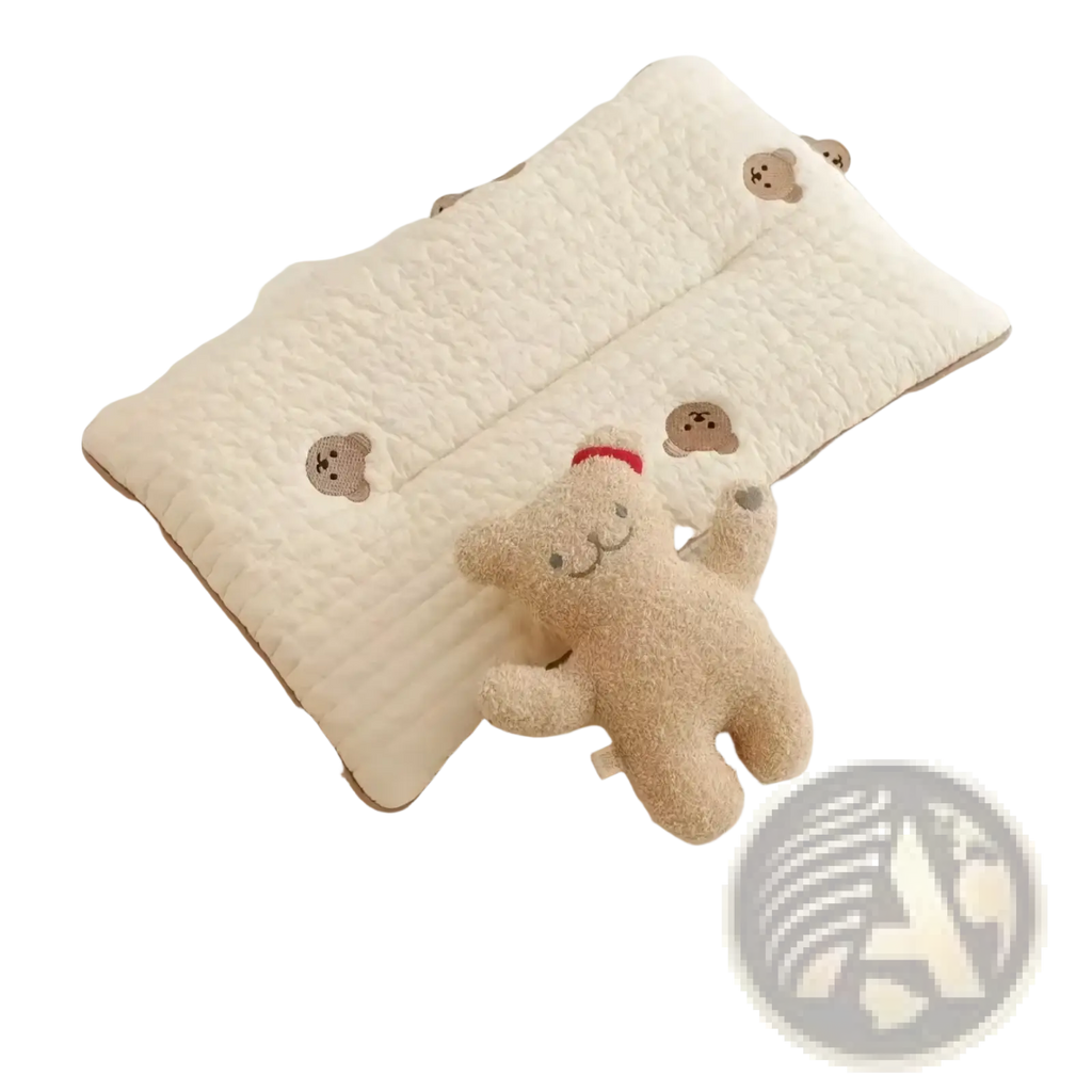 Cream-colored quilted embroidered cotton baby pillow with brown bear faces next to a plush beige teddy bear in red hat