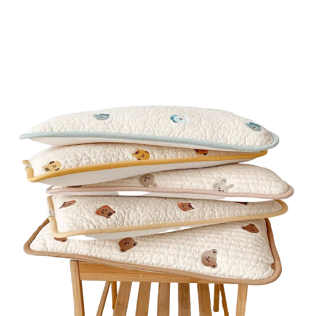 Stack of cream-colored quilted cotton baby pillows with embroidered animal faces and pastel piping