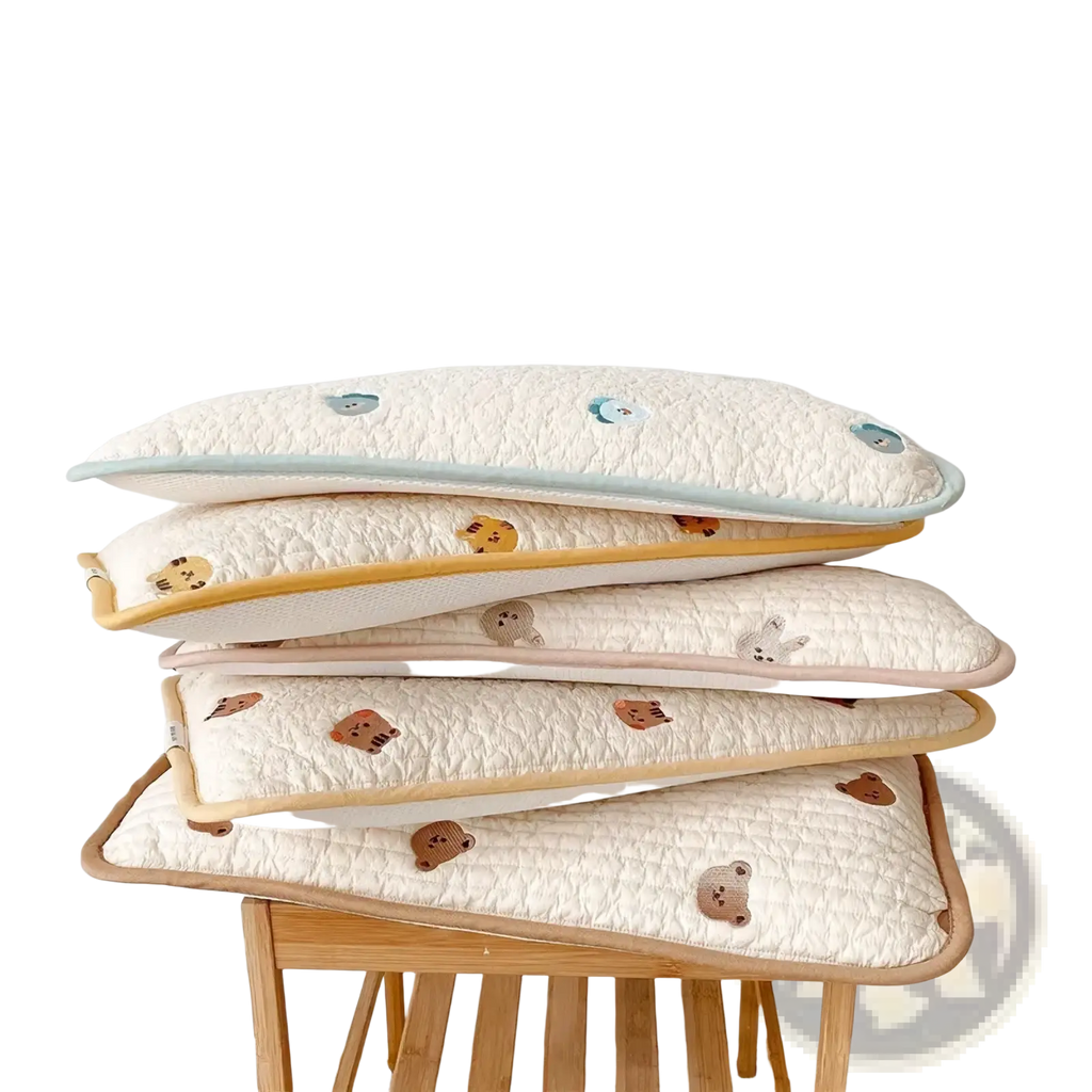 Stack of cream-colored quilted cotton baby pillows with embroidered animal faces and pastel piping