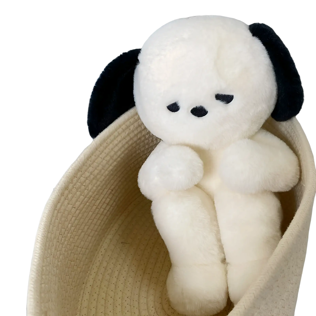 A cream-colored emo puppy plush dog with black floppy ears and embroidered face sits inside a woven basket