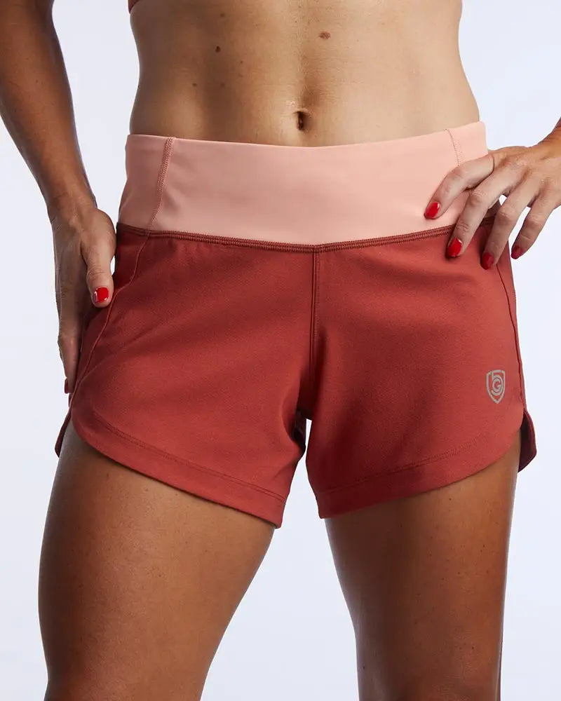 Coral-red athletic shorts with peach elastic waistband and shield logo on lower right leg for excel women’s workout