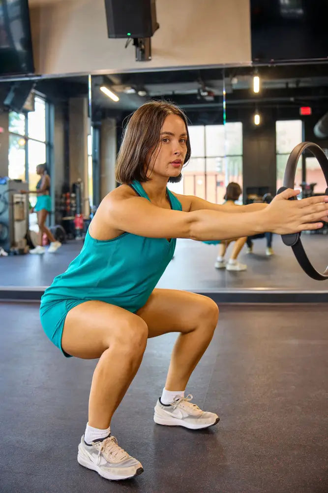 A woman performs a squat using a black pilates ring wearing excel women’s black camouflage athletic workout shorts