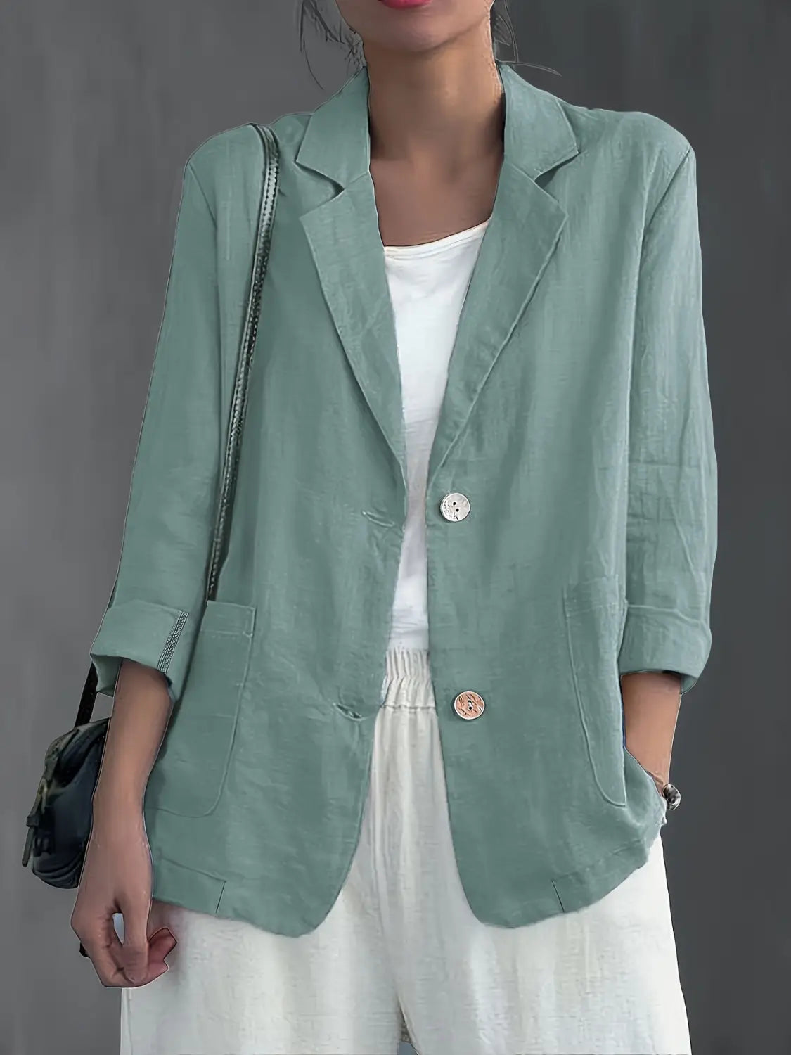 Un blazer de lino color verde salvia suave para mujer con mangas remangadas y botones circulares contrastantes, diseñado para ropa de trabajo casual.
