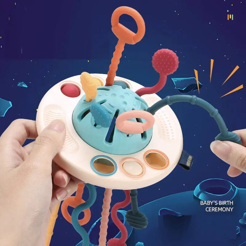 Juguete mordedor sensorial de silicona para bebé que incluye una cúpula central azul verdoso con anexos colgantes multicolores y superficies de silicona texturizadas para la estimulación táctil.