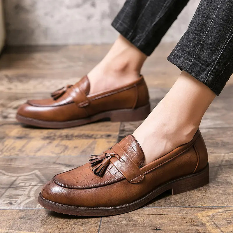 Mocasines de cuero tipo brogue con borlas para hombre en cuero texturizado color cocodrilo marrón con suela de goma, mostrados desde el lado posterior.