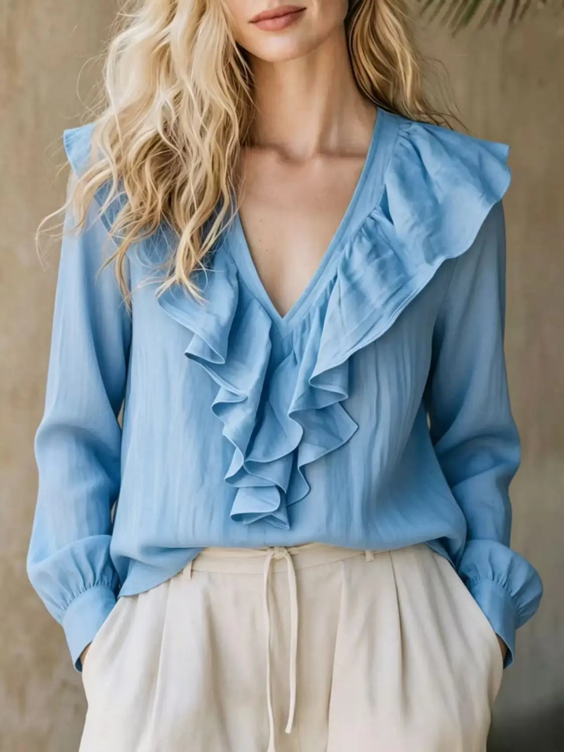 Una blusa de lino azul claro con cuello en V y manga larga, con volantes en capas a lo largo del escote, hecha de una tela ligera.