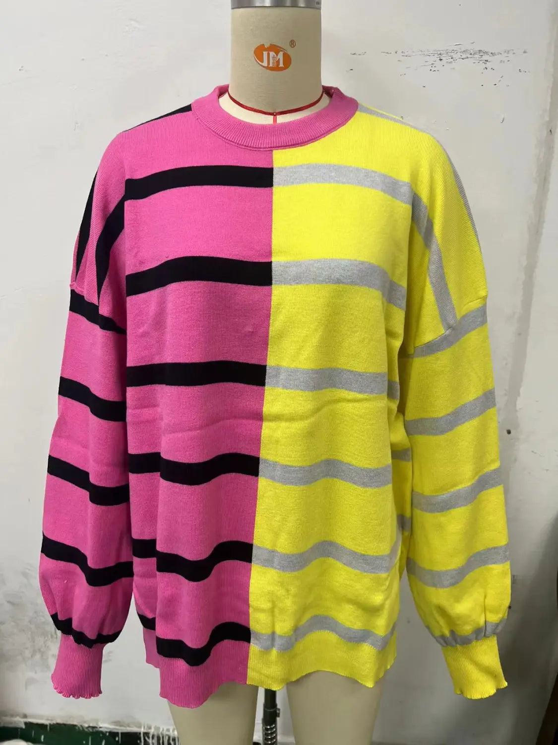 A vibrant women’s oversized split color striped knit sweater featuring bold pink and yellow sections with contrasting black and gray horizontal stripes and long sleeves with ribbed cuffs.