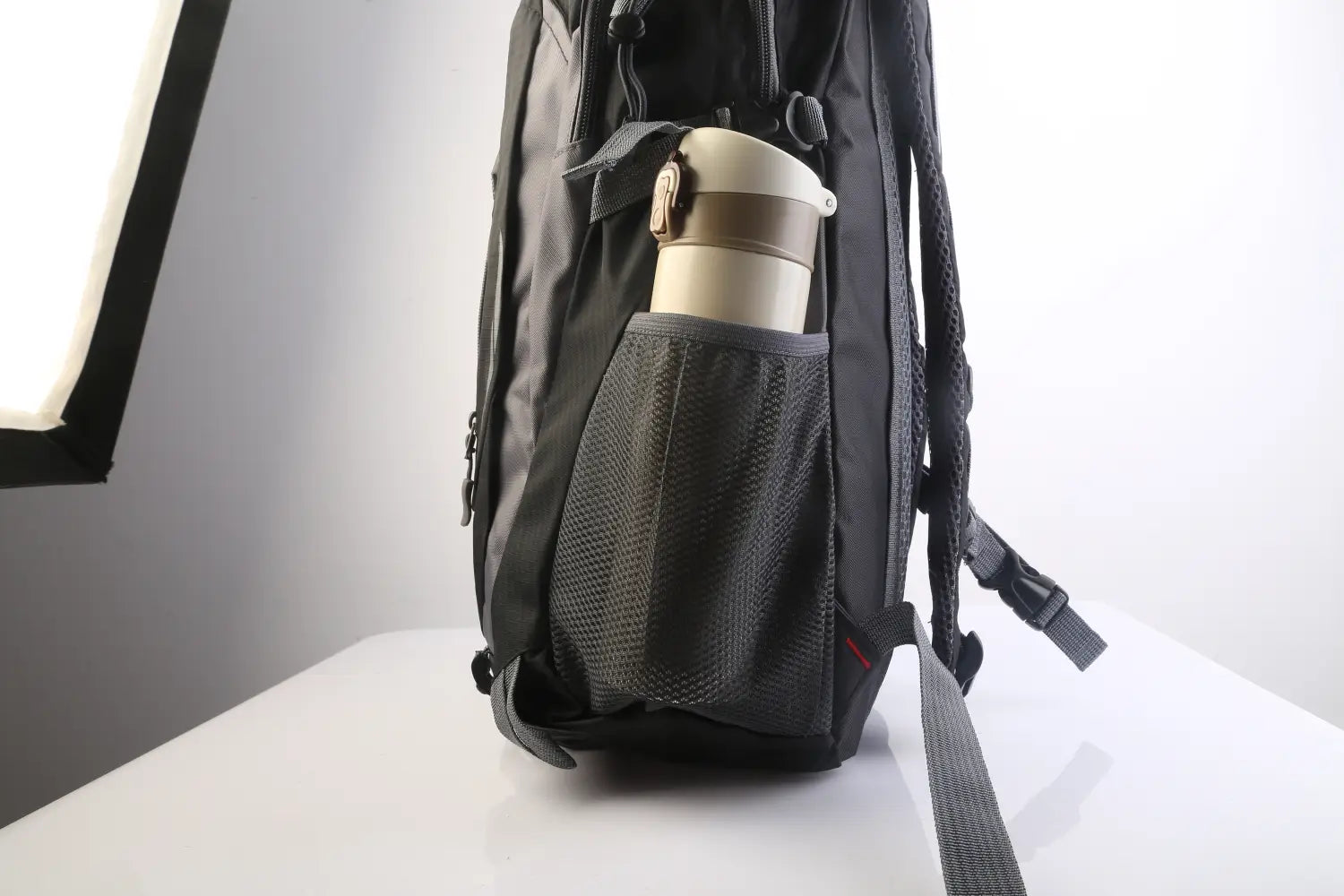 Black mesh side pocket on the back side of a 36l lightweight waterproof hiking backpack with reflective stripes holding a beige insulated travel mug with a gold-colored lid.