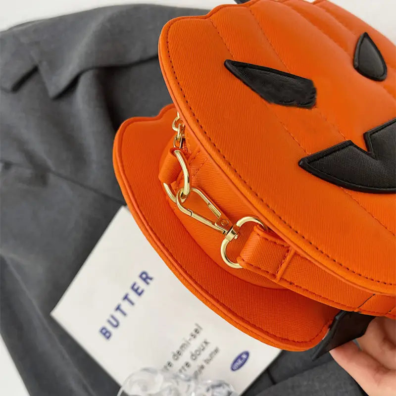 Orange pumpkin-shaped crossbody bag with black embroidered eyes and mouth featuring gold-tone hardware and a detachable bat charm.