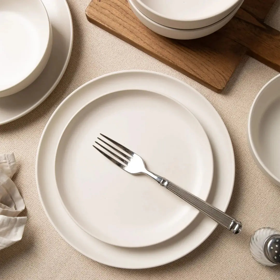 Un tenedor plateado elegante, con textura en el mango, reposa sobre un plato de cerámica blanca minimalista de un juego de vajilla de gres de 12 piezas con tazas para 3.