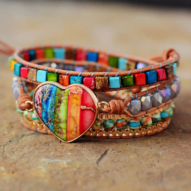 A vibrant multi-strand rainbow heart imperial stone leather wrap bracelet featuring a central rainbow-colored heart pendant with gold-tone edges and layered bands of colorful square beads faceted stones and woven leather.