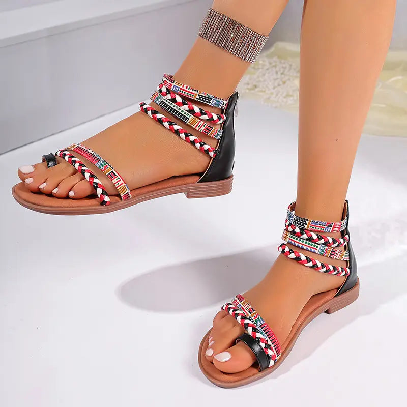 Black and tan strappy flat sandals with colorful braided ankle straps featuring a pattern of small flags in a bohemian ethnic style.