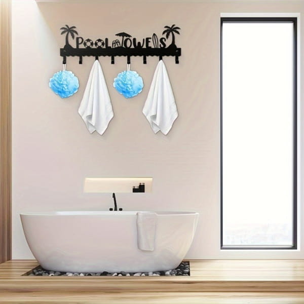Black metal pool towel rack wall mounted with eight hooks featuring palm tree and umbrella design holding two white towels and two blue bath bombs.