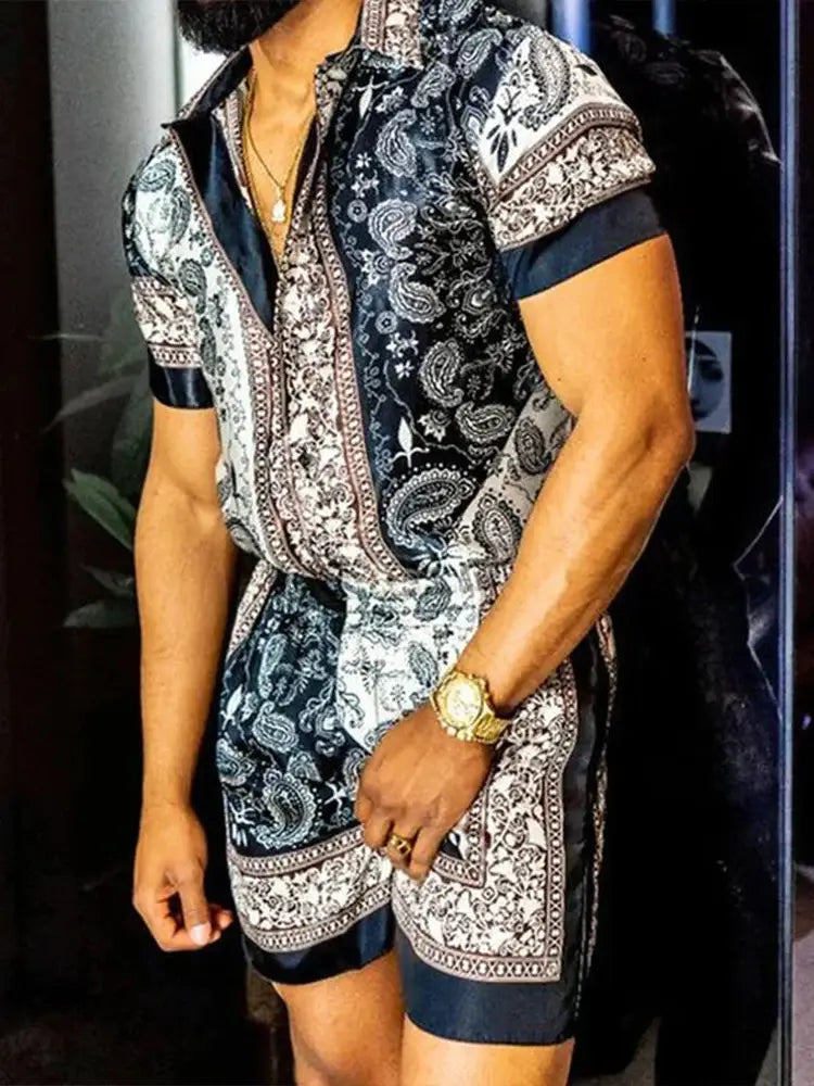 A man wearing a navy and white paisley patterned short-sleeved romper with satin trim in a men’s tropical print short sleeve shirt and shorts set.
