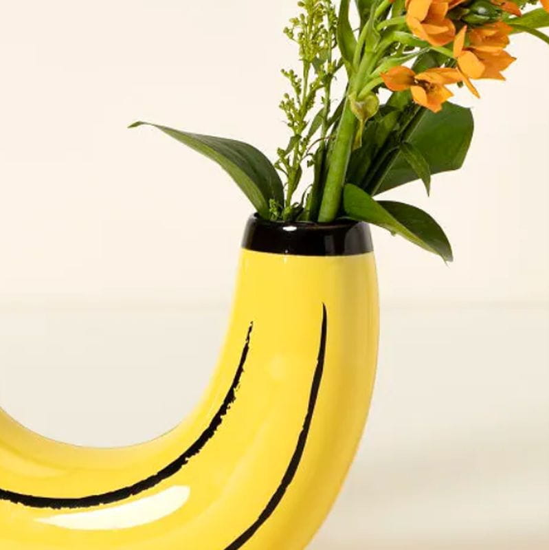 A bright yellow ceramic banana shaped vase with black curved stripes and a glossy finish holding orange flowers and green foliage for home decor.