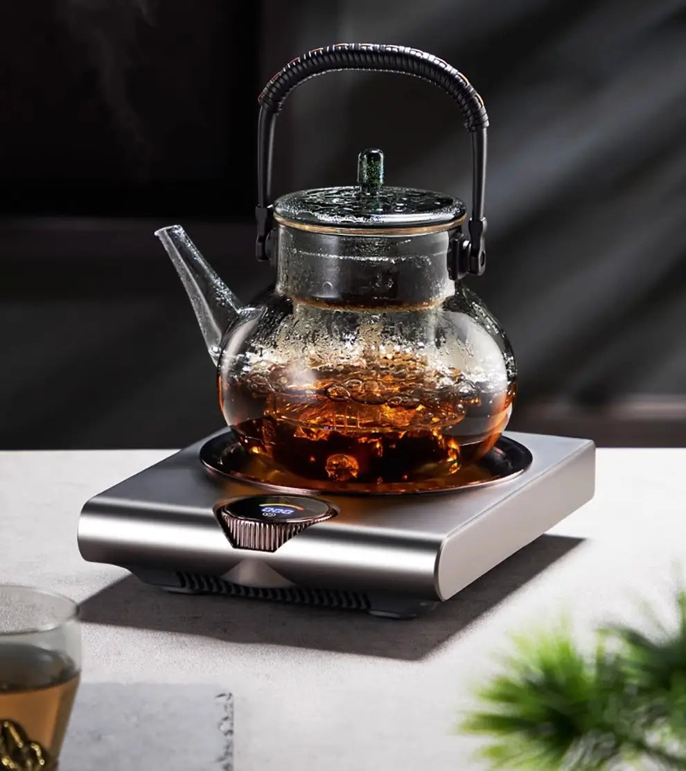 Clear glass teapot with a black metal handle and lid filled with amber tea on an electric ceramic stove 1000w tea cooker kettle.