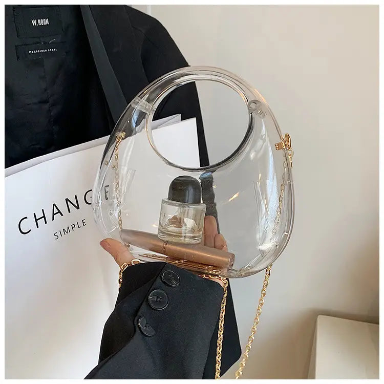 Clear acrylic handbag with a gold chain strap and metallic clutch design featuring a circular top handle.