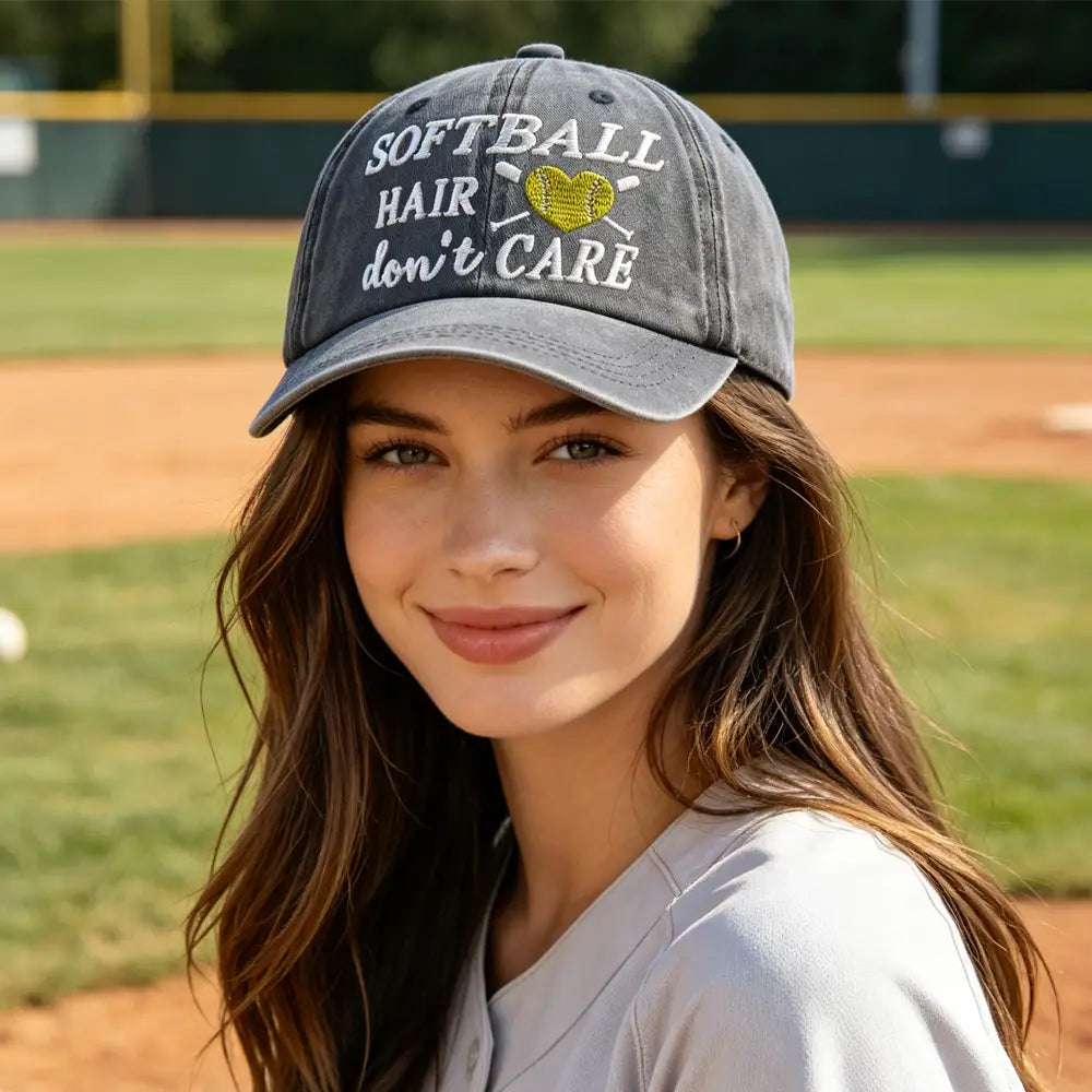 A faded gray distressed baseball cap with embroidered white text reading softball hair don’t care and a yellow heart design between crossed bats.