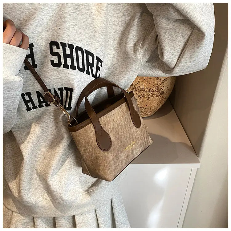 A small beige textured canvas mini tote bag with brown leather handles and trim designed as a retro bucket bag shoulder handbag.