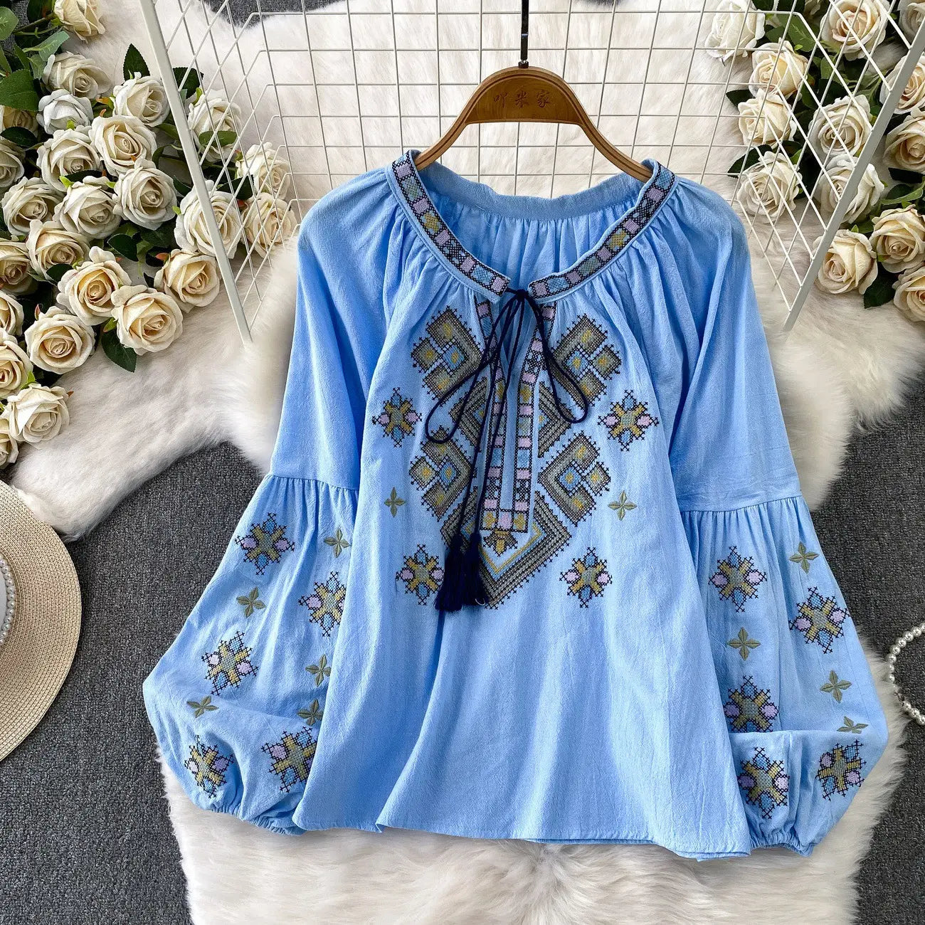 Light blue embroidered lantern sleeve tie neck blouse with geometric patterns in beige gold and purple featuring puffed sleeves and a front tie neckline.